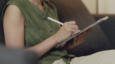 Oturma odasındaki kanepede otururken Asyalı kadın haberleri okuyor ve dijital tabletinden hisse senedi alıyor..