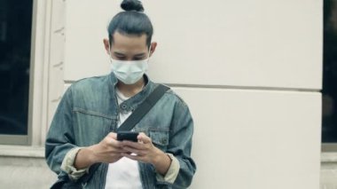 Yakışıklı Asyalı adam ameliyat maskesi takıyor ve şehir sokaklarında salgın hastalık sırasında akıllı telefon kullanıyor..