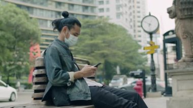 Maske takan ve akıllı telefon kullanan Asyalı adam şehrin yanındaki bankta otururken sosyal medyayı kontrol ediyor..