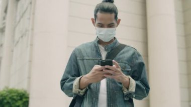 Yakışıklı Asyalı adam ameliyat maskesi takıyor ve şehir sokaklarında salgın hastalık sırasında akıllı telefon kullanıyor..