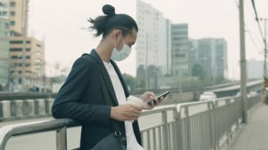 Asyalı işadamı elinde kahve fincanı olan bir yüz maskesi takıyor ve telefon kullanıyor. Şehir merkezindeki otobüs durağında Covid-19 salgını sırasında ulaşımı bekliyor..
