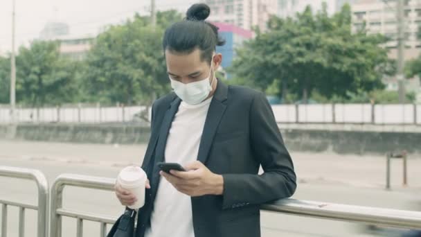 フェイスマスクをしたアジア人ビジネスマンがコーヒーカップを持っていて 流行期に街の路上輸送を待っているバスの停留所で電話を使用しています — ストック動画