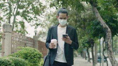 Yakışıklı Asyalı bir işadamı ameliyat maskesi takıyor ve şehir sokaklarında yürürken akıllı telefon kullanıyor..