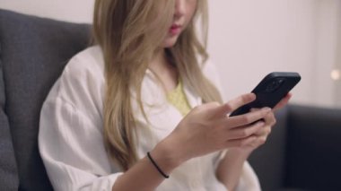 Beyaz tişörtlü Asyalı kadın kanepede oturuyor ve akıllı telefon kullanıyor oturma odasında sosyal medyada geziniyor..