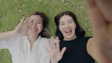 İki genç bayan arkadaş akıllı telefonlarda kendi portrelerini çekiyorlar ve yeşil çimenlerde uzanmış tatilin tadını çıkarıyorlar..