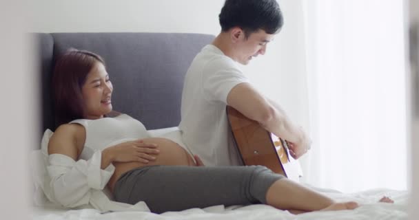 アジアの夫と妊娠中の妻は一緒にベッドルームで時間を過ごし 夫は家で生まれていない赤ん坊のためにギターの歌を歌う — ストック動画