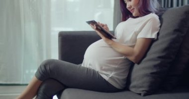 Güzel Asyalı hamile kadın evdeki oturma odasında bir koltukta otururken karnını okşuyor..