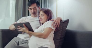 Yeni doğmuş bebeğinin ultrason fotoğrafına bakan hamile bir karısı olan mutlu beyaz bir koca. Tablet üzerinde ve evdeki yatak odasında internetten alışveriş yapıyor..