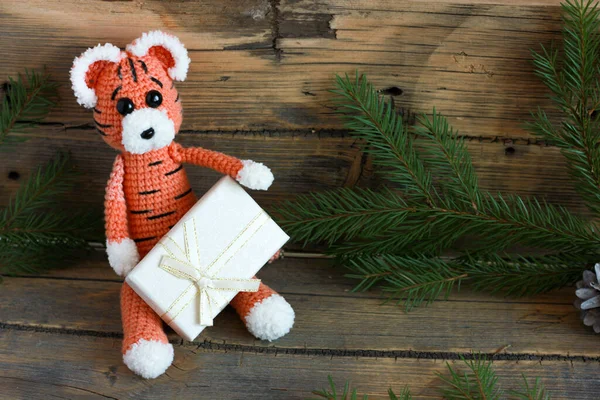 crocheted tiger sitting with a gift on a wooden background with branches of a christmas tree in the background.