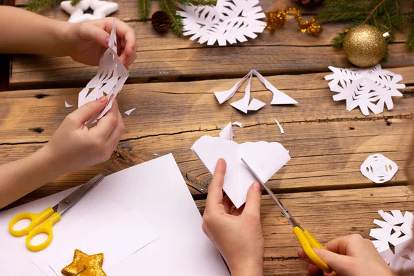 Kinderhände schneiden zu Hause Schneeflocken aus Papier auf Holzgrund Stockfoto