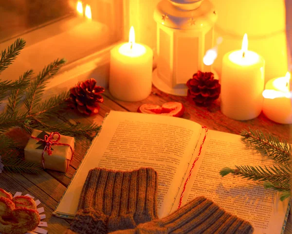 Kazağın kolları kitabın üzerinde duruyor. Evde, pencerenin üzerinde hediye olarak mumlar olan samimi bir Noel akşamının kompozisyonu.