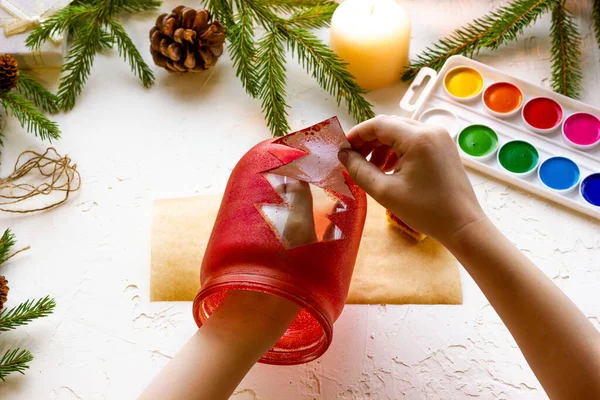Les mains des garçons font un chandelier à partir d'un pot en verre peint avec de la peinture rouge avec une silhouette d'un arbre de Noël, instructions étape par étape, étape 7. — Photo
