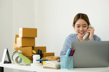 Genç Asyalı kadın işyeri sahibi masanın üzerinde mutlu online satış işi, dizüstü bilgisayarı kullan, müşterilerden emir al, notlar al ve teslimat için postayla ayarlamaları yap..