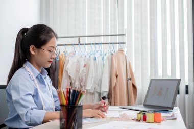 Tekstil aksesuarları ve girişimci tasarımcı çizimleriyle genç ve güzel Asyalı moda terzisi stüdyoda yaratıcı fikirler için dizüstü bilgisayarı olan parlak renklerle dolu..