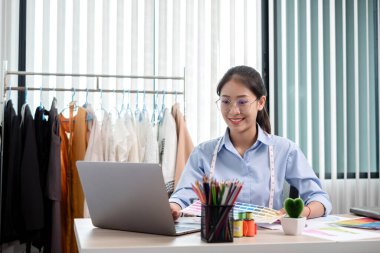 Tekstil aksesuarları ve girişimci tasarımcı çizimleriyle genç ve güzel Asyalı moda terzisi stüdyoda yaratıcı fikirler için dizüstü bilgisayarı olan parlak renklerle dolu..