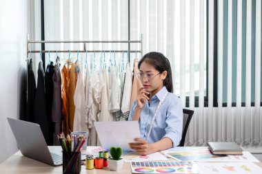 Tekstil aksesuarları ve girişimci tasarımcı çizimleriyle genç ve güzel Asyalı moda terzisi stüdyoda yaratıcı fikirler için dizüstü bilgisayarı olan parlak renklerle dolu..