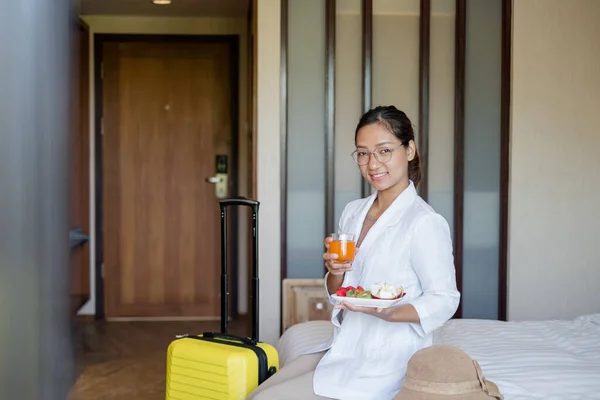 An Asian businesswoman sits on a bed in a hotel room, exhausted from a business trip, ready to receive a fruit snack from the hotel, arranged for VIP guests.