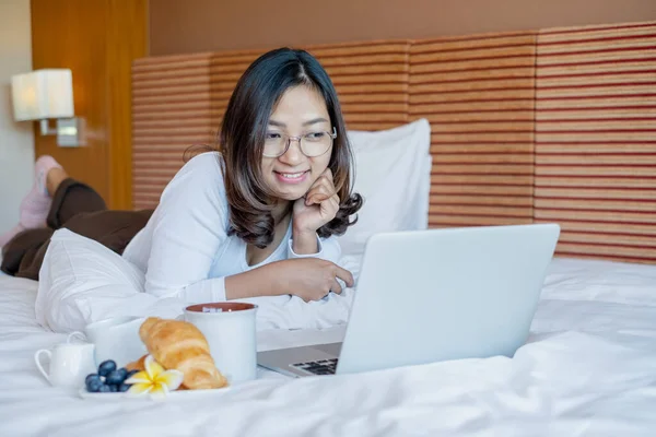 Young Asian women smiling happily at freelance work, working on a notebook while relaxing in bed with snacks and fruit in a hotel room. Vacation and relaxation.