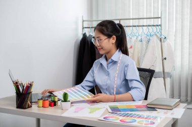 Tekstil aksesuarları ve girişimci tasarımcı çizimleriyle genç ve güzel Asyalı moda terzisi stüdyoda yaratıcı fikirler için dizüstü bilgisayarı olan parlak renklerle dolu..