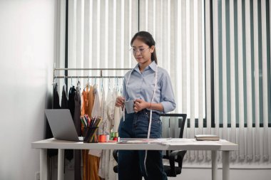 Tekstil aksesuarları ve girişimci tasarımcı çizimleriyle genç ve güzel Asyalı moda terzisi stüdyoda yaratıcı fikirler için dizüstü bilgisayarı olan parlak renklerle dolu..