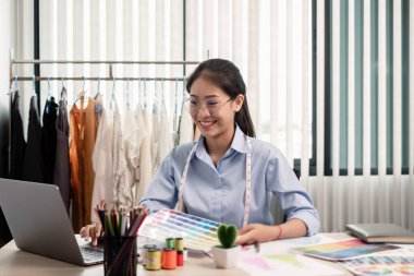 Tekstil aksesuarları ve girişimci tasarımcı çizimleriyle genç ve güzel Asyalı moda terzisi stüdyoda yaratıcı fikirler için dizüstü bilgisayarı olan parlak renklerle dolu..