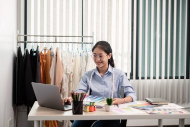 Tekstil aksesuarları ve girişimci tasarımcı çizimleriyle genç ve güzel Asyalı moda terzisi stüdyoda yaratıcı fikirler için dizüstü bilgisayarı olan parlak renklerle dolu..