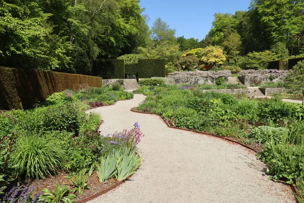 A irregular curved gravel path runs between flowerbeds of abundant perennial flowers in the formal gardens of an English country house on the edge of Dartmoor National park in Devon, England