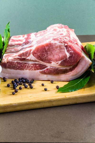 Pork loin or back joint with ingredients for dry curing into bacon, salt, sugar, peppercorns, juniper berries and bay leaves, portrait and centered, on wooden chopping board elevated view.