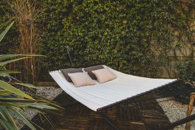 Özel bir ev ya da şehir otelinin bahçesinde iki kişilik büyük boy modern bir hamağın yakın çekimi. Arka planda yasemin bitkisi duvarı, rahatlatıcı bir yer..