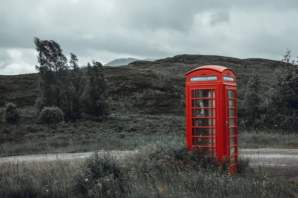 British vintage red phone booth in the middle of nowhere, along a country road in Scotland. overcast cloudy day, rain drizzle. Contrast between technology and nature, red and green.