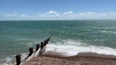 Brighton yakınlarındaki bir İngiliz güney kıyısı kasabasında, denizin beyaz köpüğüyle güçlü dalgaların üzerindeki statik kamera. Çakıl taşları ve kayalardan oluşan kumsal manzarası, açık mavi deniz ve gökyüzü, güneşli bir gün. İngiltere 'de yaz günü.