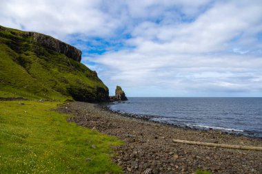 Wide angle views of Talisker Bay, Isle of Skye, Scotland, with its rocky beach, black stones, green fields, and a waterfall at the end of beach. Scattered clouds on blue sky, summer scottish weather.