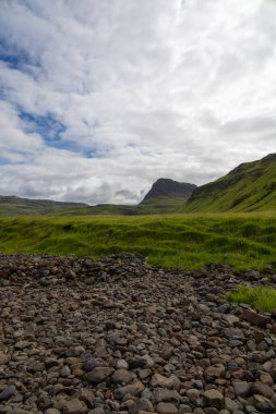 Wide angle views of Talisker Bay, Isle of Skye, Scotland, with its rocky beach, black stones, green fields, and a waterfall at the end of beach. Scattered clouds on blue sky, summer scottish weather.
