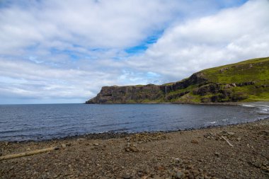Wide angle views of Talisker Bay, Isle of Skye, Scotland, with its rocky beach, black stones, green fields, and a waterfall at the end of beach. Scattered clouds on blue sky, summer scottish weather.