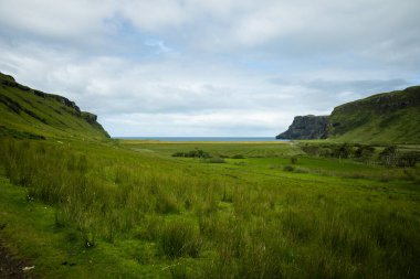 Wide angle views of Talisker Bay, Isle of Skye, Scotland, with its rocky beach, black stones, green fields, and a waterfall at the end of beach. Scattered clouds on blue sky, summer scottish weather.