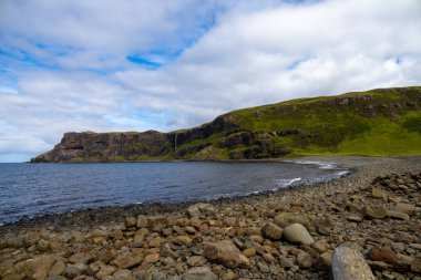 Wide angle views of Talisker Bay, Isle of Skye, Scotland, with its rocky beach, black stones, green fields, and a waterfall at the end of beach. Scattered clouds on blue sky, summer scottish weather.