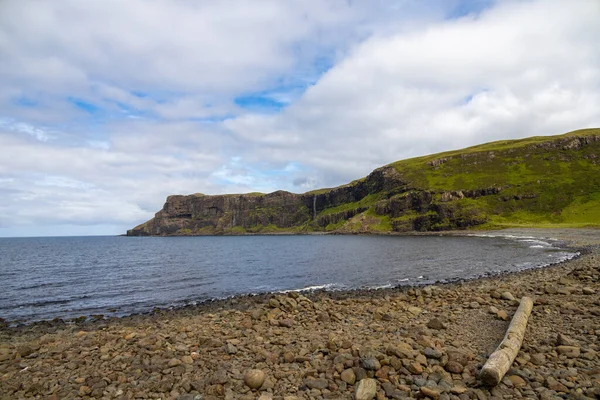 Wide angle views of Talisker Bay, Isle of Skye, Scotland, with its rocky beach, black stones, green fields, and a waterfall at the end of beach. Scattered clouds on blue sky, summer scottish weather.