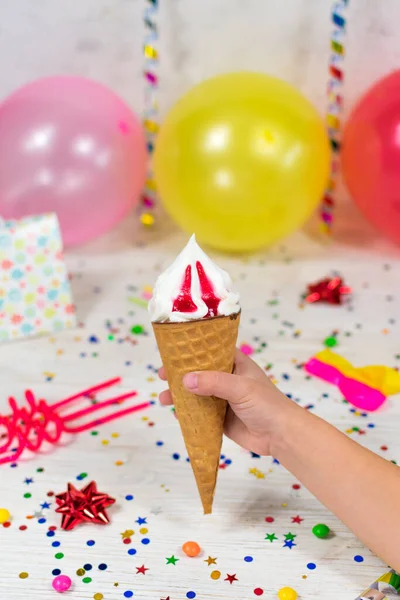 Cone de sorvete em uma mão de crianças em um fundo de aniversário com decoração colorida e doces com um fundo borrado. Foco seletivo. — Fotografia de Stock