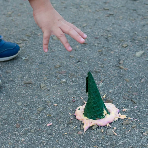 An upset childs hand reaches for the ice cream that has fallen on the pavement. Bright ice cream in a green cone fell on the asphalt. — Stock Photo, Image