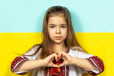 Ulusal Ukrayna elbiseli, nakışlı gömlekli bir kız Ukrayna bayrağının arka planına yakın plan çekilmiş bir Ukrayna sevgisi işareti olarak kalp işareti gösteriyor. Ukrayna 'daki savaşı durdurun. Şey...