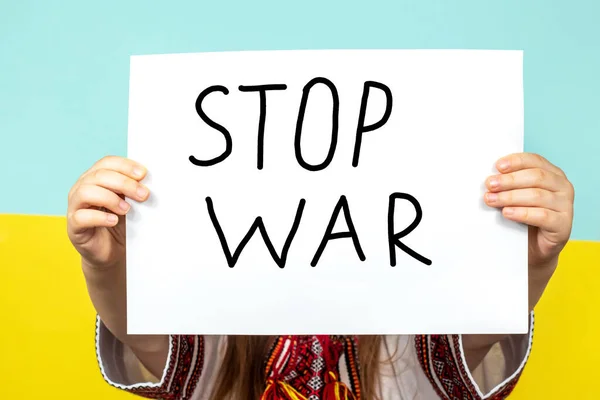 El primer plano de STOP WAR se sostiene en las manos de una niña sobre el fondo de la bandera ucraniana. Detener la guerra en Ucrania. El concepto de paz en Ucrania. — Foto de Stock