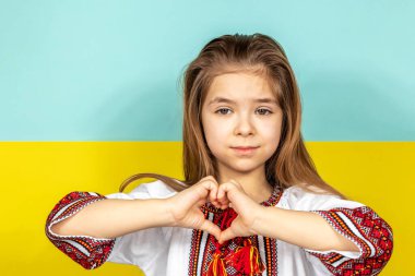 Ulusal Ukrayna elbiseli, nakışlı gömlekli bir kız Ukrayna bayrağının arka planına yakın plan çekilmiş bir Ukrayna sevgisi işareti olarak kalp işareti gösteriyor. Ukrayna 'daki savaşı durdurun. Şey...