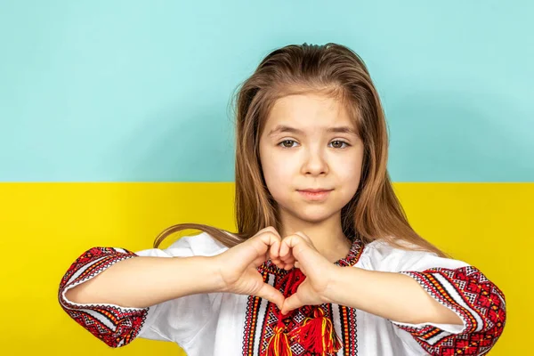 Uma menina de roupas ucranianas nacionais, uma camisa bordada, mostra um sinal de coração como um sinal de amor pela Ucrânia, close-up contra o fundo da bandeira ucraniana. Parem a guerra na Ucrânia. O — Fotografia de Stock