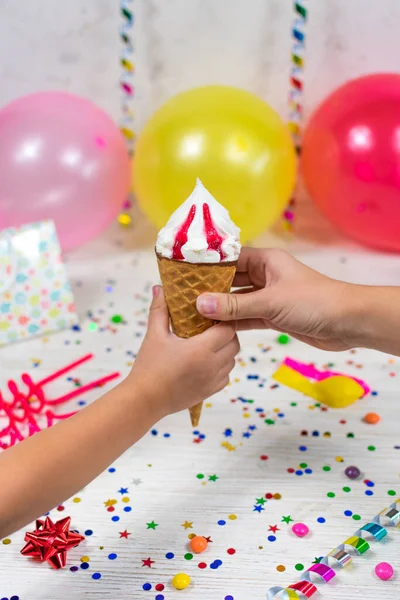 Moms hand passes an ice cream cone to the childs hand on a birthday background with colorful decor and sweets with blurred background. Selective focus