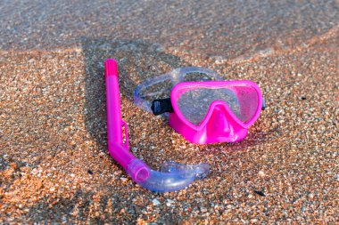 Pink swimming mask and diving snorkel lie on the sand near the water.