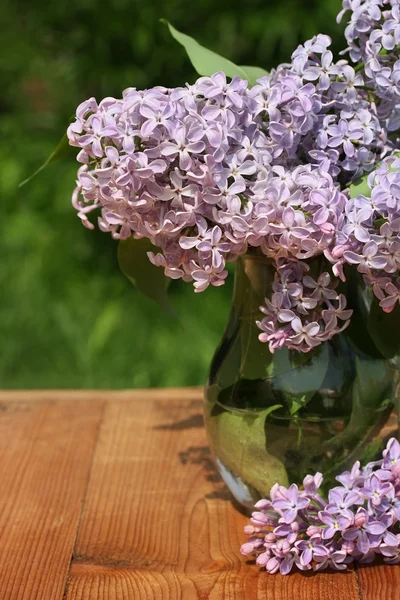 Bouquet de lilas dans une cruche en verre sur une table en bois contre les verts — Photo