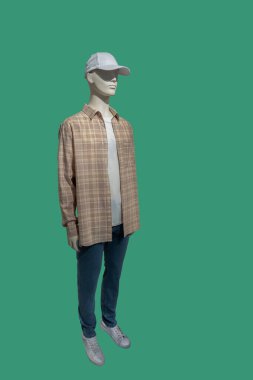 Bir erkek vitrin mankeni, düğmesiz uzun kollu ekose gömlek ve yeşil arka planda izole edilmiş mavi kot pantolon giyer.