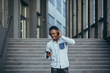 Genç Afrikalı öğrenci akıllı telefondan müzik dinliyor. Büyük kulaklıklar kullanıyor. Uygulamanın kolaylığından gülümsüyor.