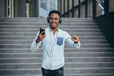 Genç Afrikalı öğrenci akıllı telefondan müzik dinliyor. Büyük kulaklıklar kullanıyor. Uygulamanın kolaylığından gülümsüyor.