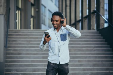 Genç Afrikalı öğrenci akıllı telefondan müzik dinliyor. Büyük kulaklıklar kullanıyor. Uygulamanın kolaylığından gülümsüyor.